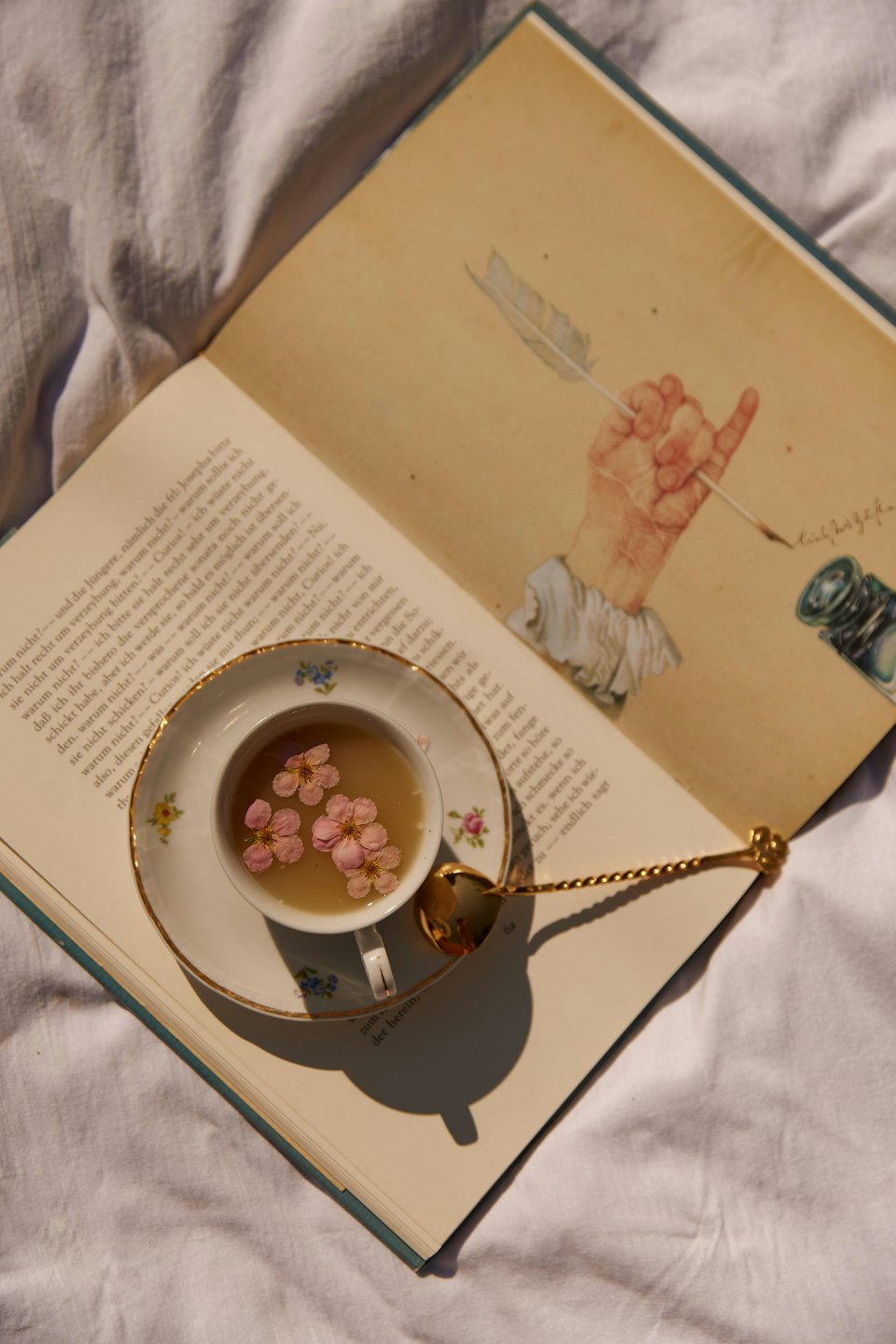An open antique book and tea rest on white bedding.